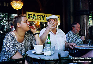 An older man wearing a white cap stretches his hand to count while a chubby younger woman listens at Budapest's Müvés Café in Hungary.