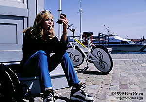 An attractive blonde girl is adjusting her make up in front of her parked free city bicycle while a ferry boat passes by in the port of Copenhagen, Denmark.