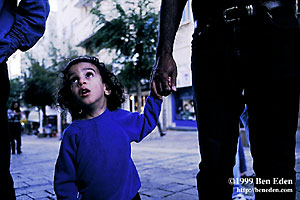 A Jewish boy wearing a kippa over black, curly hair, in a blue sweatshirt, looks up while holding his father's hand at the Midrahove pedestrian zone in Jerusalem, Israel