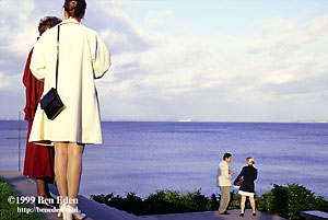 Two elegant couples chat outside, lit by golden hour light, during a museum opening in Humlebk, Denmark, while Sweden's Helsingsborg is visible across the sea.