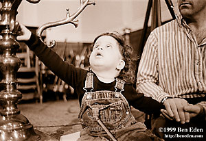 A little Jewish boy with long, curly hair, in lace-up jeans pants, reaches at a big antique bronze Menorah while his father holds his other hand at the children's Chanukah (Hanukah) party at the Prague Jewish Community.