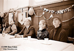 A girl sitting at a table watching the action surrounded by her grandparents at a Chanukah (Hanukah) children's party held in Prague, Czech Republic Jewish Community's eating hall.