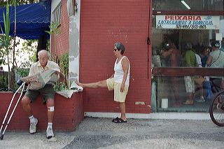 Female brazilian senior stretches on the sidewalk while an invalide reads the newspaper next to a fish store on Copacabana beach, Rio de Janeiro, Brazil