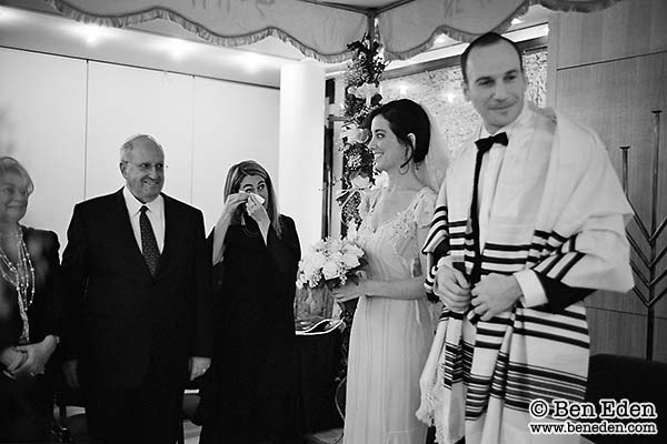 Jewish bride and groom stand under the Chuppah watched by family